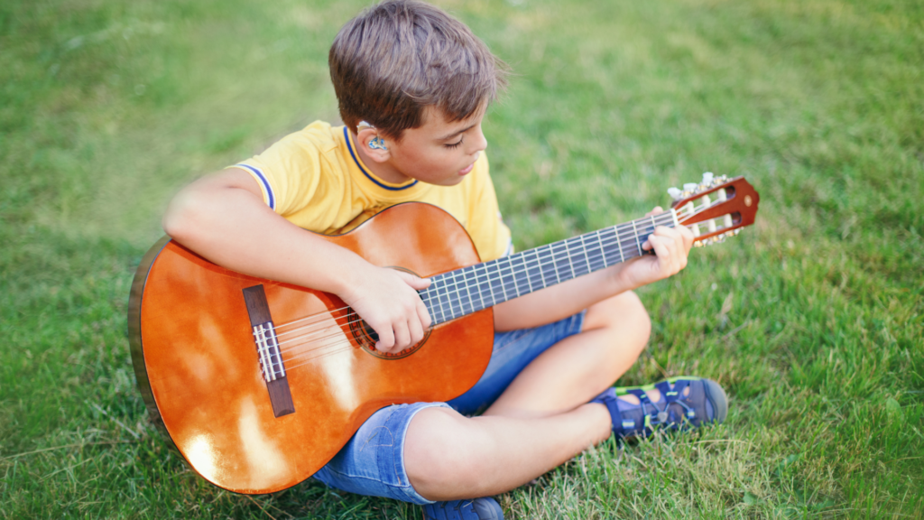 Enfant ou jeune garçon atteint de problèmes auditifs portant un embout de prothèse auditive sur mesure pour son appareil auditif jouant de la guitare dans l'herbe. 